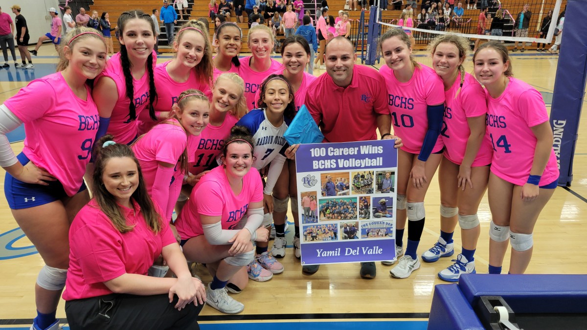 Barron Collier volleyball coach Yamil Del Valle holds sign celebrating his 100th career, which also happened come as his team won its 13th consecutive district championship with a win over Naples.
